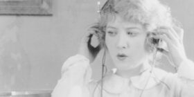 historic photograph shows women sitting next to radio listening to radio broadcast by holding headphones to ears.