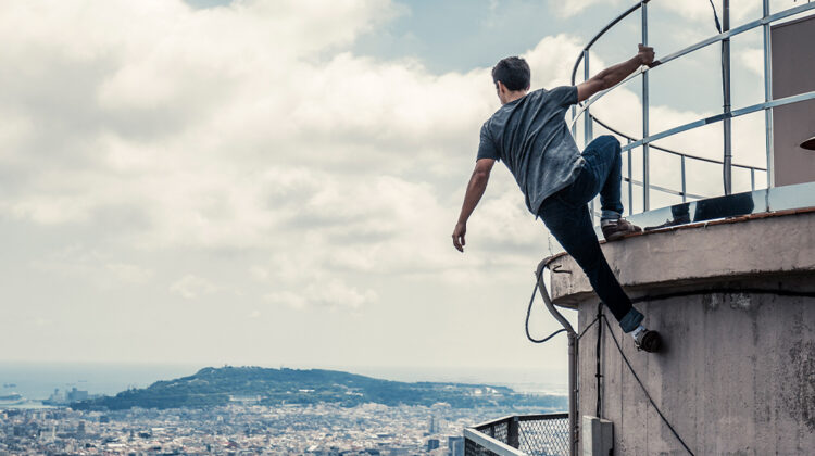 Man practicing parkour and hanging from the side of tall structure looking out at the city