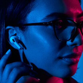 Young female model in eyeglasses and modern true wireless earbuds listening to audio in studio with neon lights